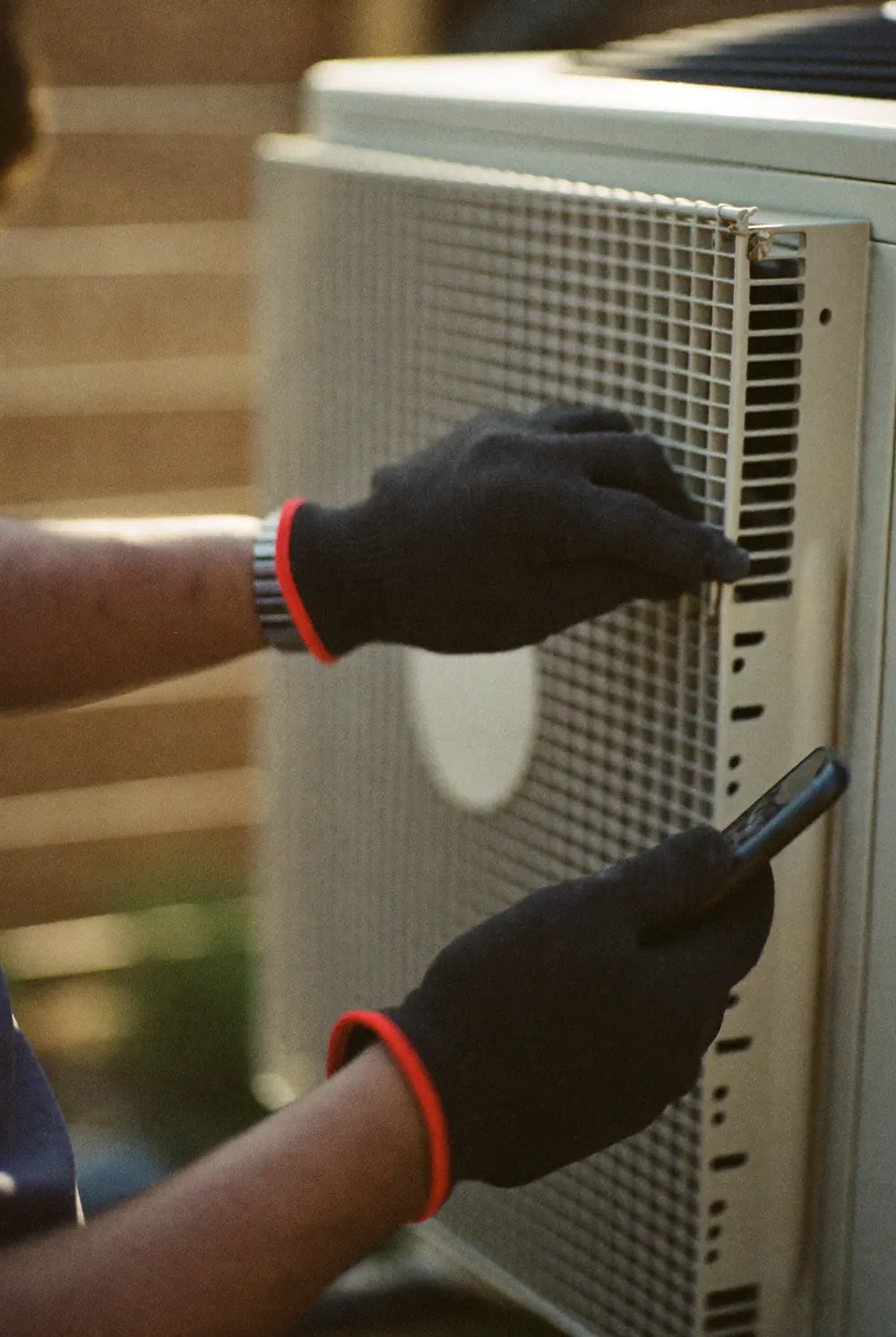 A technician in work gloves opens the grille of an outdoor air conditioner unit while holding a phone