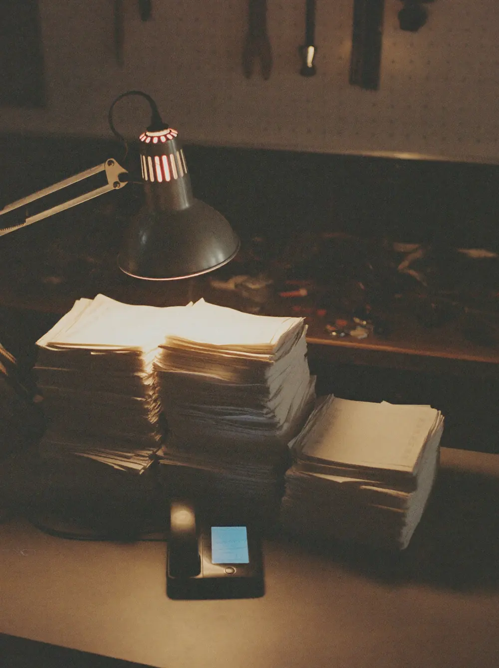 A work lamp lights stacks of paper invoices on a workshop desk at night beside a phone