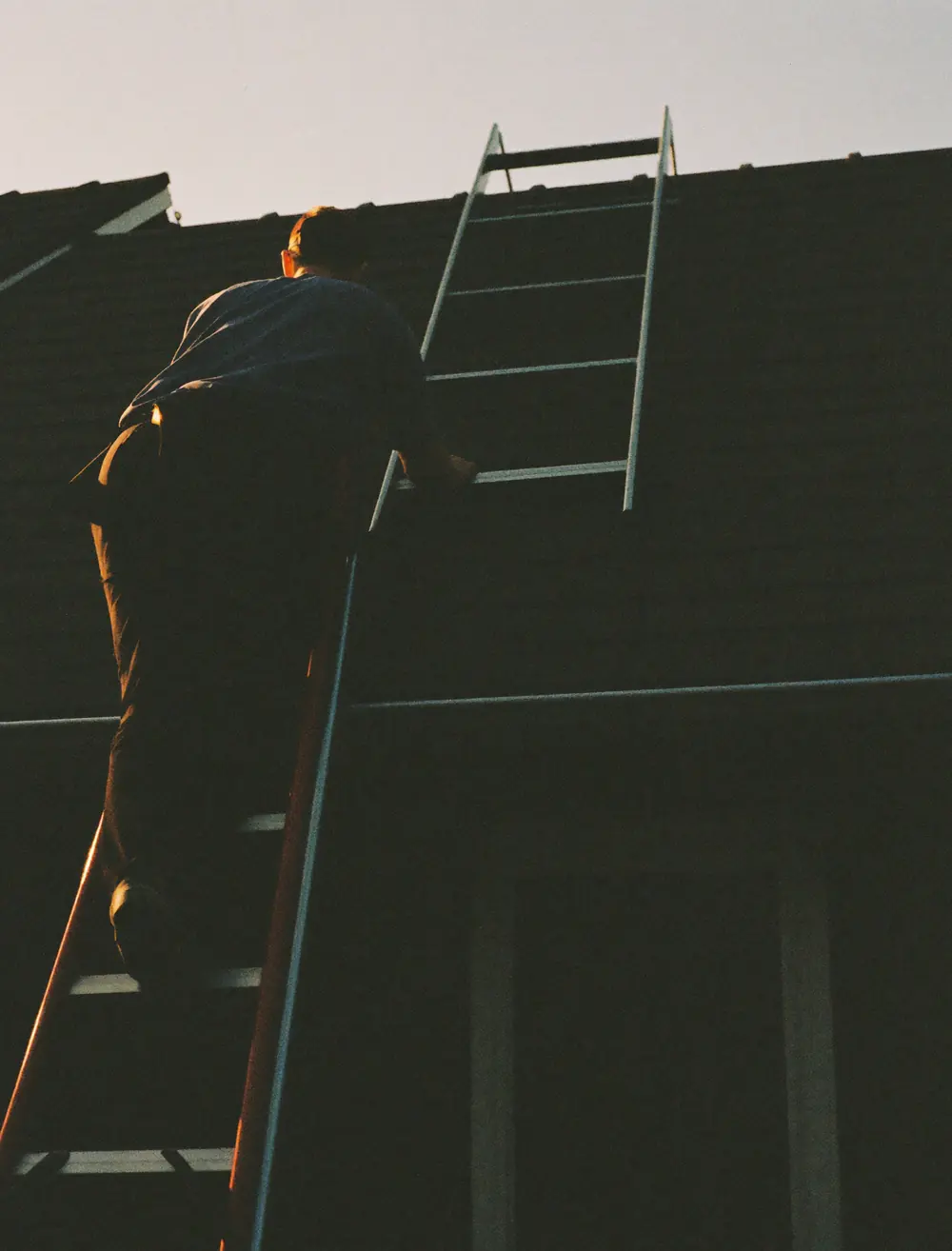 A roofer climbing a ladder onto a roof at dusk, seen from behind