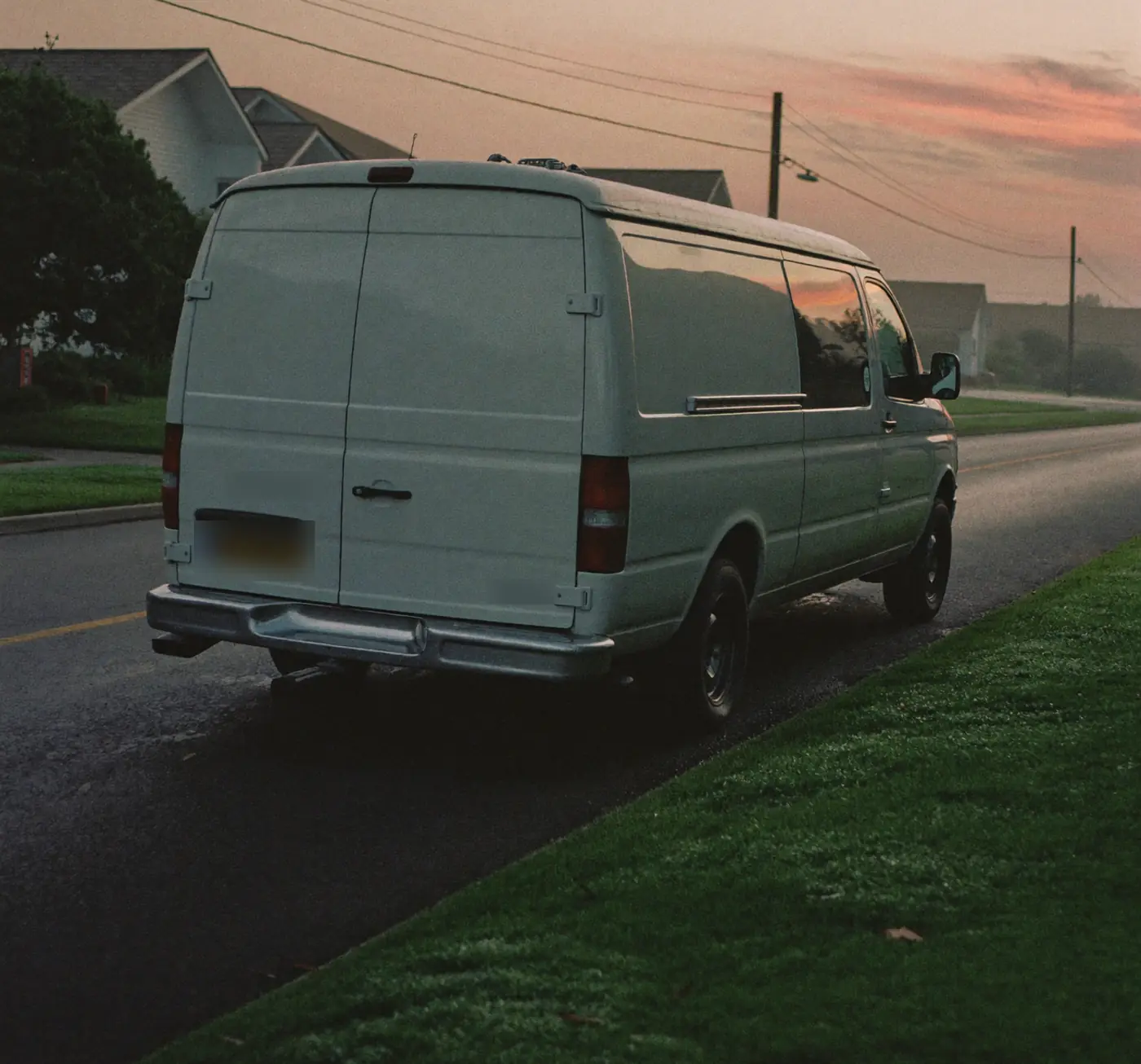 A white service van parked on a quiet suburban street at dawn under a pink sky